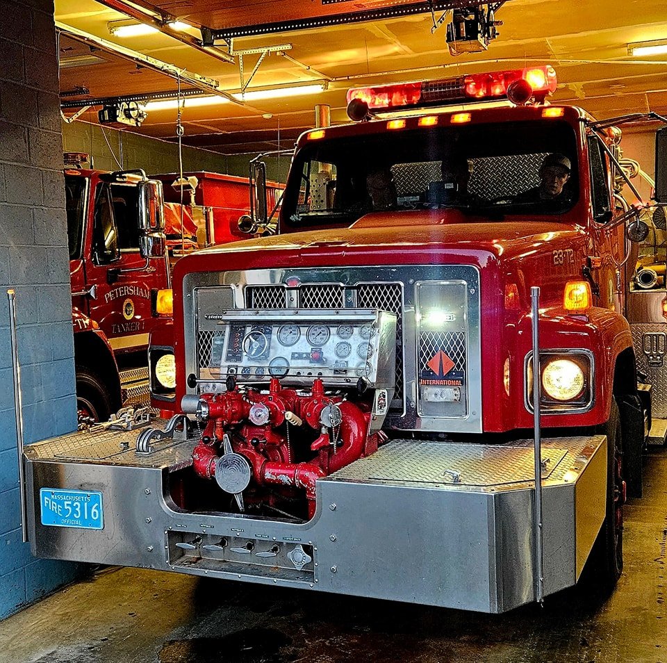 Two red Petersham Volunteer Fire Department engines parked in the station bay; the rear truck is lettered Petersham Tanker 1.